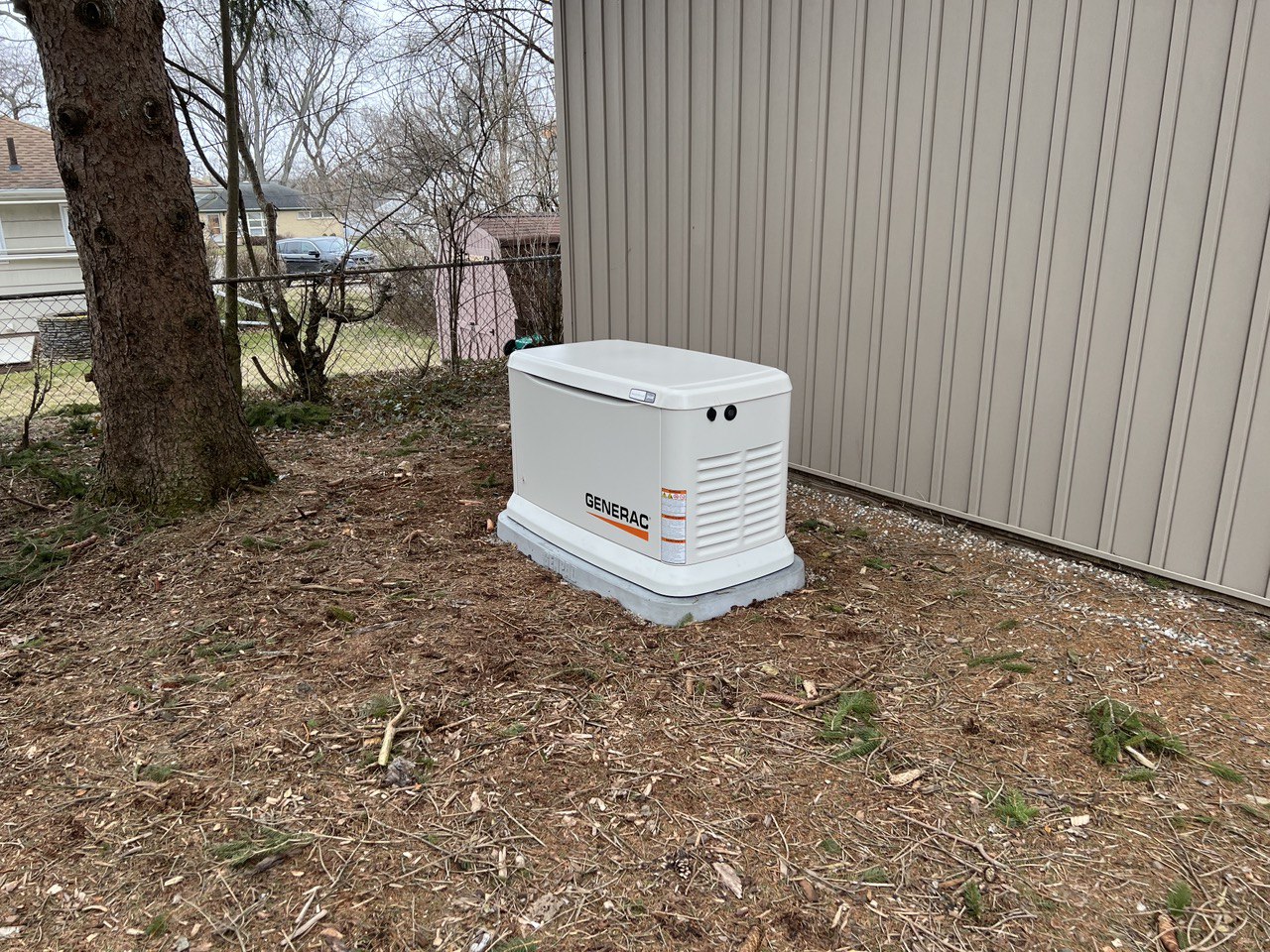 Generac Guardian 26kw generator sitting on a concrete pad next to a tan shed under a pine tree